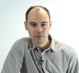 Man stares at the camera, against a light blue backdrop. He is wearing a grey shirt. 